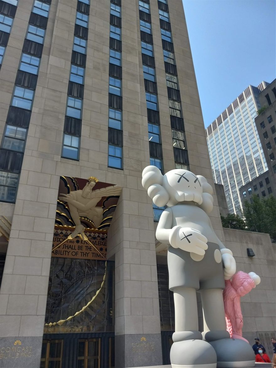 Walking past 30 Rockefeller Center with a KAWS sculpture and a quote from the Bible; 